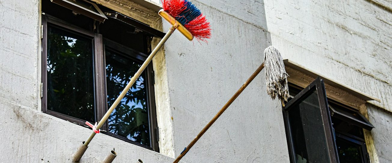Farbiger Besen und Mopp hängen an der Wand eines Gebäudes mit offenen Fenstern.