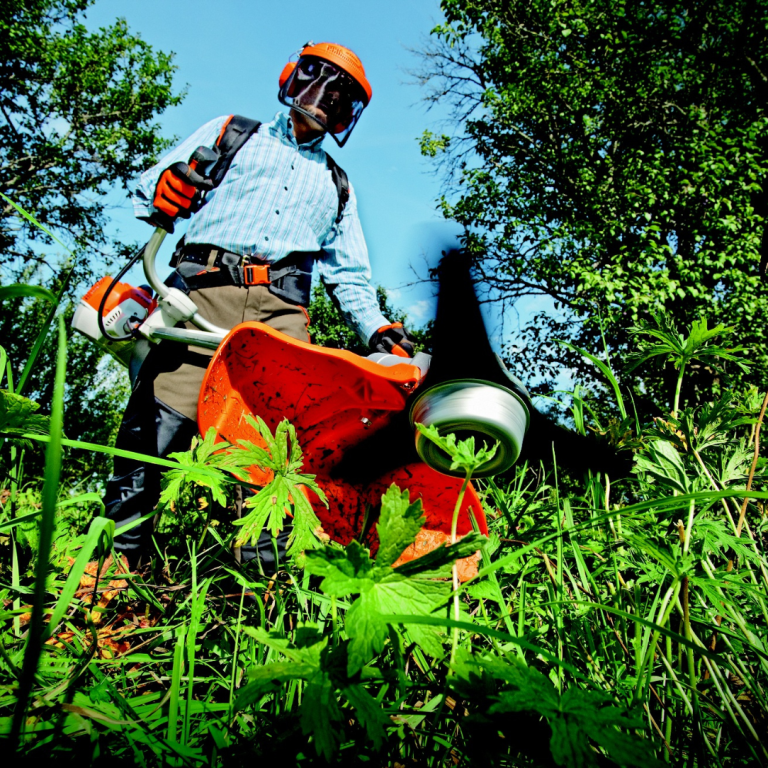 Mann mit einem Freischneider in grünem Gelände, umgeben von Pflanzen und Bäumen.